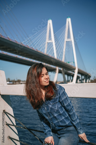 A brunette girl with long hair in a blue shirt stands against the background of a cable stayed bridge on the embankment