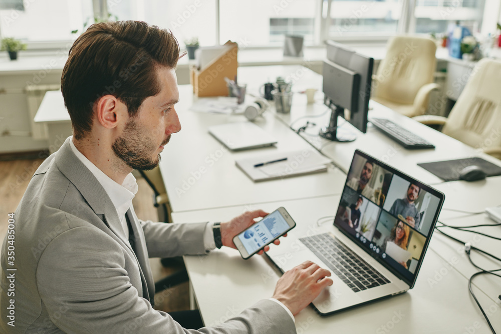 Obraz premium Young bearded businessman working alone in office while connecting colleagues via video chat on laptop during coronavirus epidemic