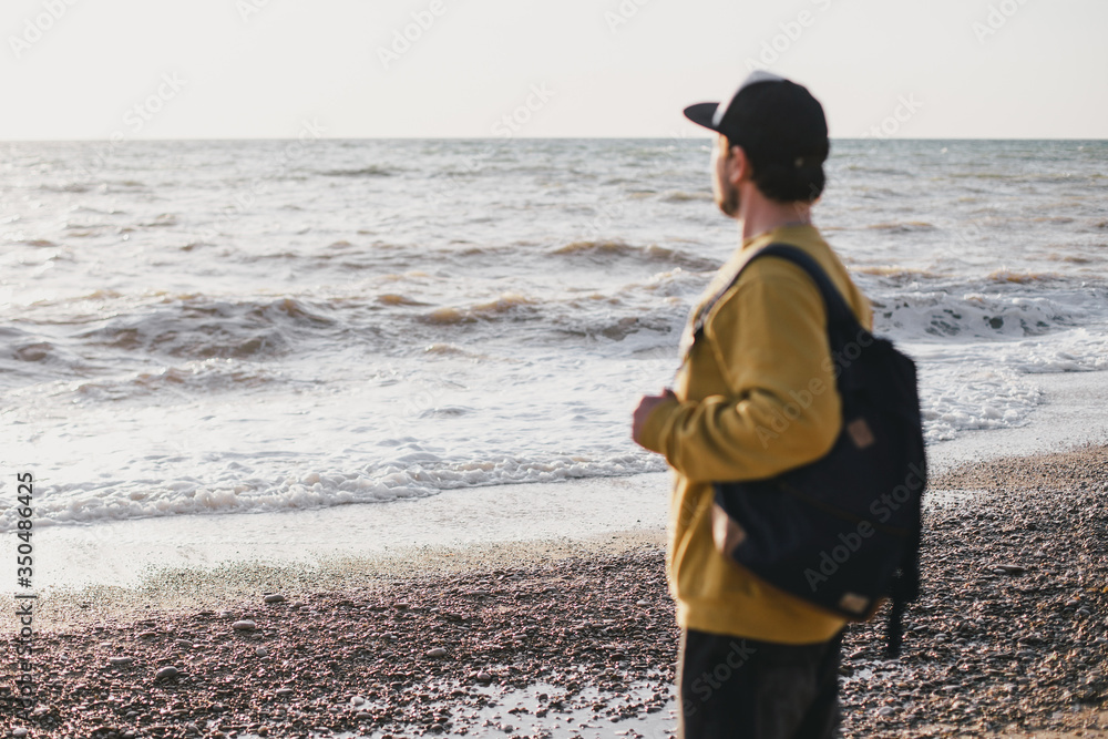 Young man traveler with beard wearing backpack exploring empty beach near ocean or sea.