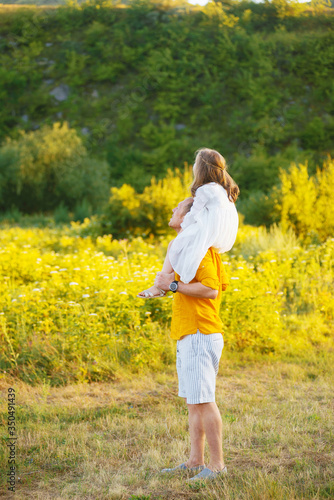 Father is holding on his shoulders his little girl in white dress looking far away.