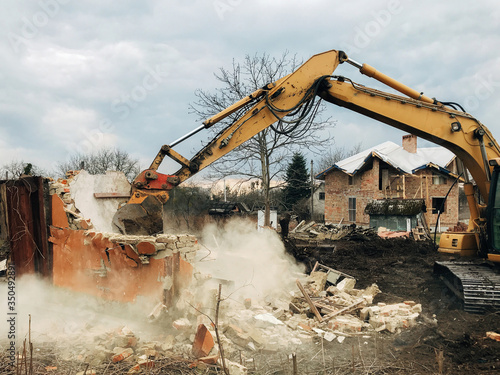 House crushing and collapse. Excavator destroying brick house on land in countryside. Bulldozer clearing land from old bricks and concrete from walls with dirt and trash. Ruining house
