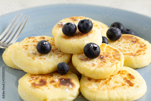 Cheese pancakes close-up on a blue ceramic plate decorated with blueberries and icing sugar with a fork on the left