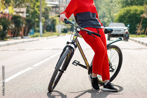 Sports lifestyle. A woman in red sportswear, posing leaning on a Bicycle. In the background, an empty road and street. Eco-friendly transport concept. Close up of legs