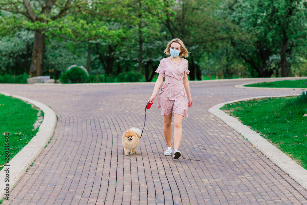 Fototapeta premium Young woman and red spitz with a medical mask on her face on nature on a spring day. Coronavirus pandemic
