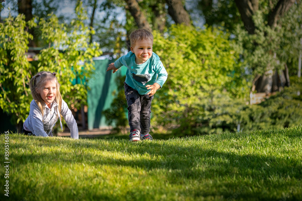 Fototapeta premium Two cute adorable playful caucasian blond children siblings little boy girl enjoy having fun playing together at home backtard green grass lawn at summer day. Brother sister happy childhood concept