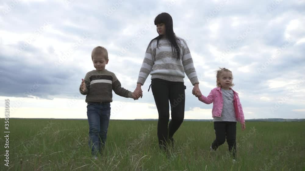 A young mother with her children walking on the green grass, holding hands. A happy family enjoys nature, walking, and relationships. The concept of motherhood, parenting. A young mother with her children walking on the green grass, holding hands. A happy family enjoys nature, walking, and relationships. The concept of motherhood, parenting.