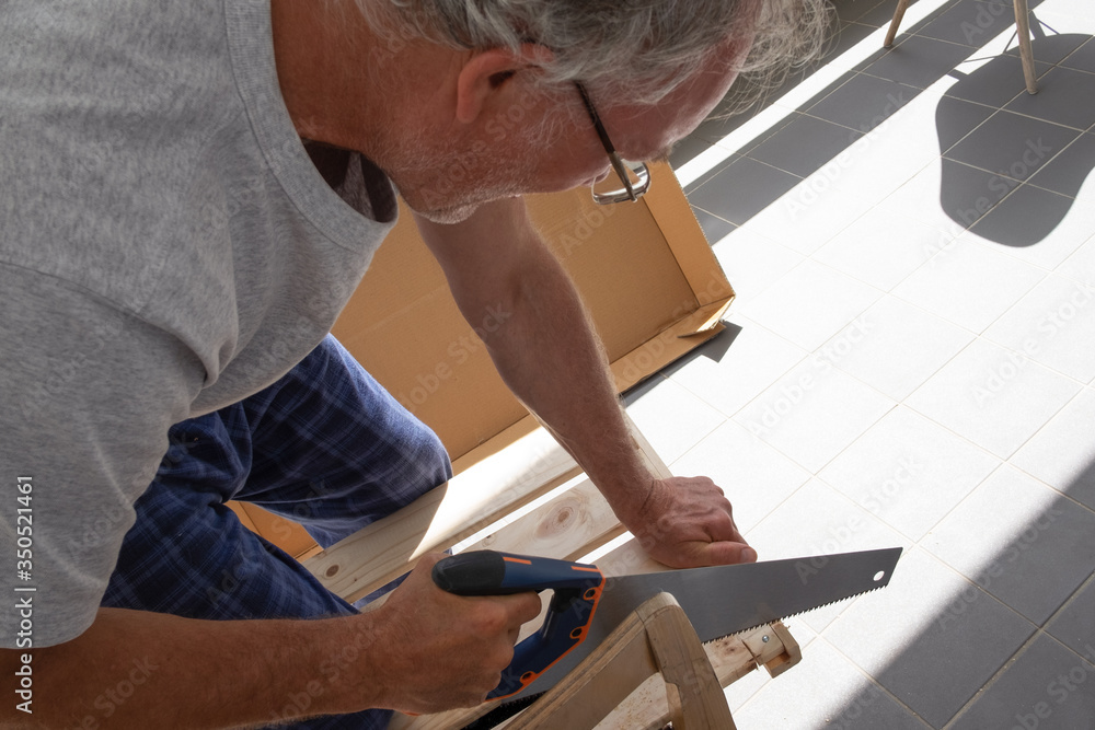 Mature man wearing home clothes, cutting wooden planks with saw, doing carpentry work. Male household chores or craft hobby concept