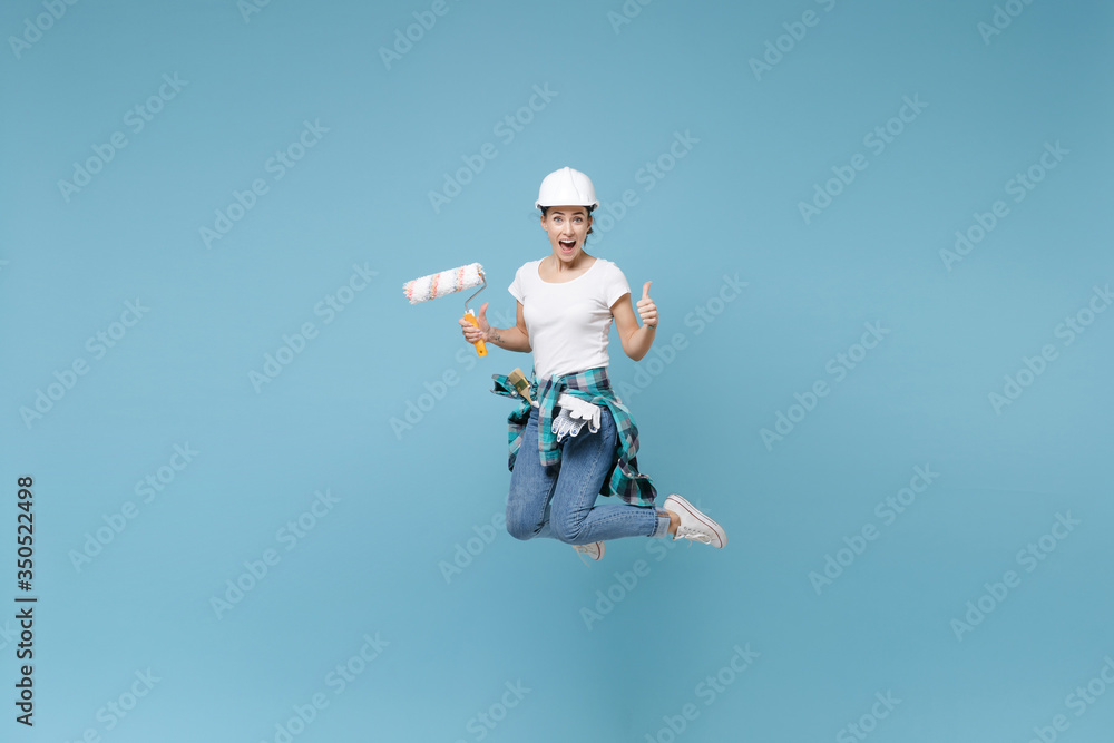 Excited young woman in protective helmet hardhat jump with paint roller isolated on blue wall background. Instruments accessories for renovation apartment room. Repair home concept. Showing thumb up. Excited young woman in protective helmet hardhat jump with paint roller isolated on blue wall background. Instruments accessories for renovation apartment room. Repair home concept. Showing thumb up.