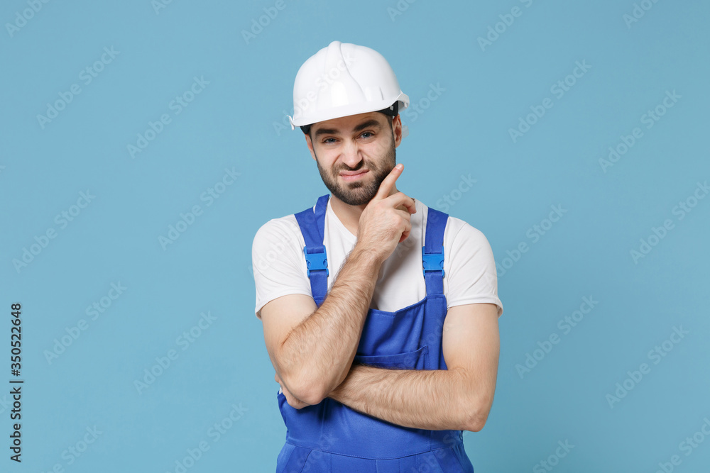 Confused young man in coveralls protective helmet hardhat isolated on pastel blue background. Instruments accessories for renovation apartment room. Repair home concept. Put hand prop up on chin. Confused young man in coveralls protective helmet hardhat isolated on pastel blue background. Instruments accessories for renovation apartment room. Repair home concept. Put hand prop up on chin.