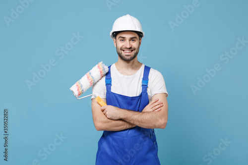 Smiling man in coveralls protective helmet hardhat hold paint roller isolated on blue background. Instruments accessories for renovation apartment room. Repair home concept. Holding hands crossed. photo
