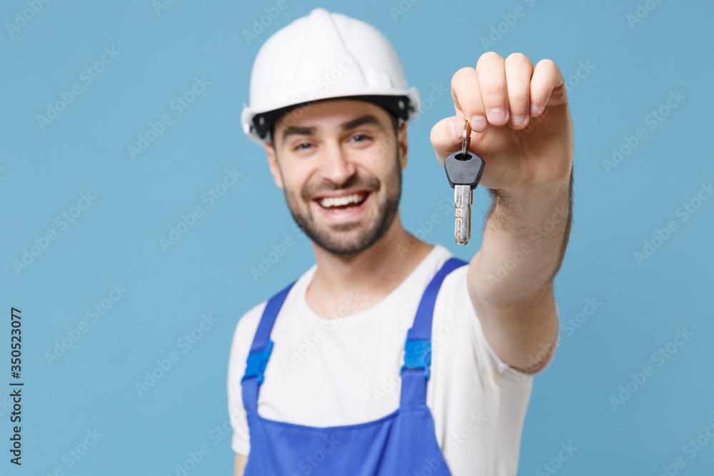 Cheerful young man in coveralls protective helmet hardhat hold keys isolated on pastel blue wall background studio portrait. Instruments accessories for renovation apartment room. Repair home concept.