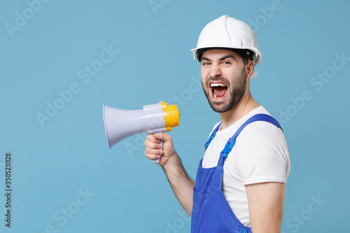Side view of angry young man in coveralls protective helmet hardhat scream in megaphone isolated on blue background studio. Instruments accessories for renovation apartment room. Repair home concept.
