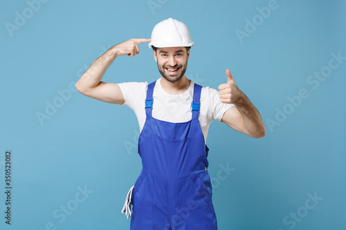Smiling man in coveralls pointing index finger on protective helmet hardhat isolated on blue background . Instruments accessories for renovation apartment room. Repair home concept. Showing thumb up.