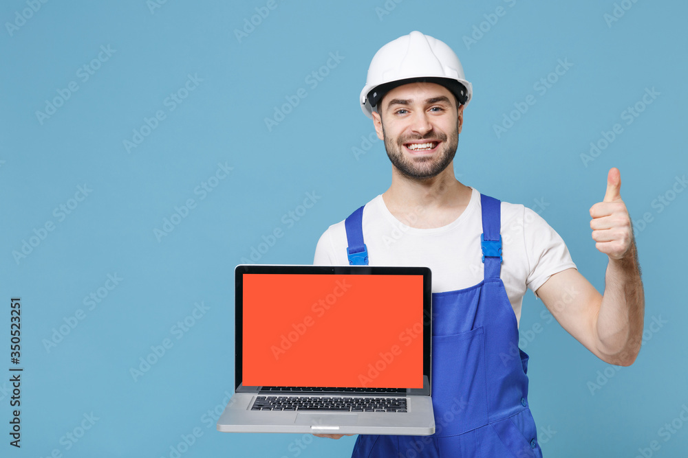 Smiling man in coveralls hardhat hold laptop computer with blank empty screen isolated on blue background. Instruments accessories renovation apartment room. Repair home concept. Showing thumb up. Smiling man in coveralls hardhat hold laptop computer with blank empty screen isolated on blue background. Instruments accessories renovation apartment room. Repair home concept. Showing thumb up.