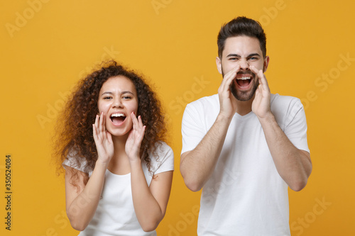 Crazy couple two friends european guy african american girl in white t-shirts posing isolated on yellow background. People lifestyle concept. Mock up copy space. Scream with hands gesture near mouth.