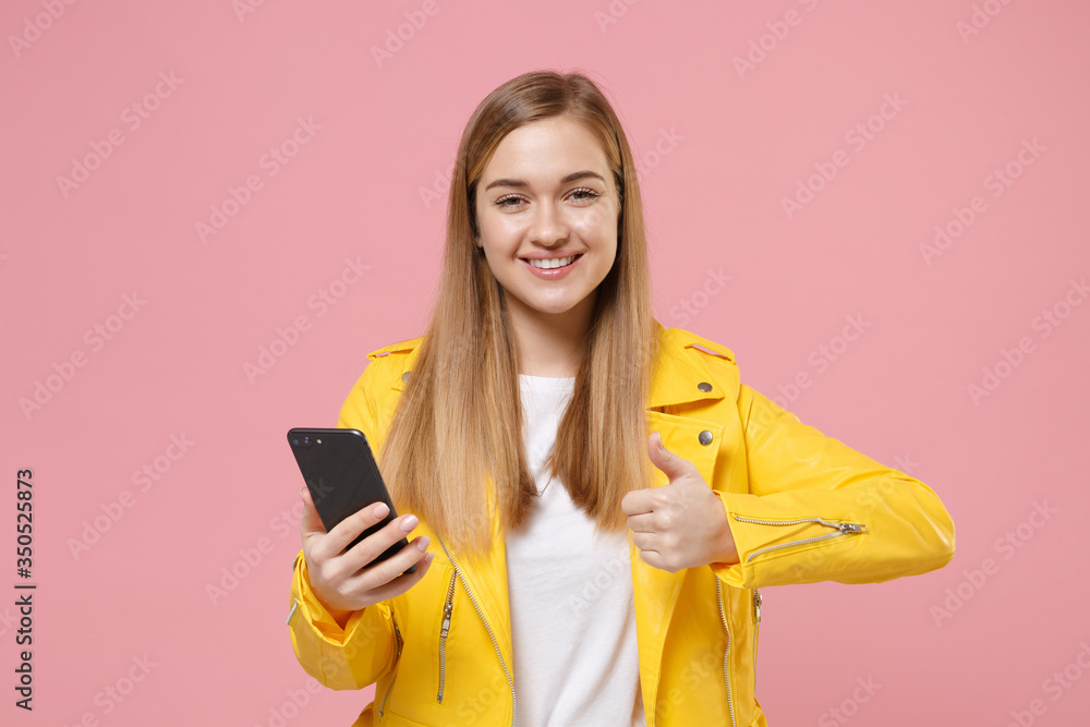 Smiling young woman girl in yellow leather jacket posing isolated on pastel pink wall background studio portrait. People lifestyle concept. Mock up copy space. Hold mobile phone, showing thumb up. Smiling young woman girl in yellow leather jacket posing isolated on pastel pink wall background studio portrait. People lifestyle concept. Mock up copy space. Hold mobile phone, showing thumb up.