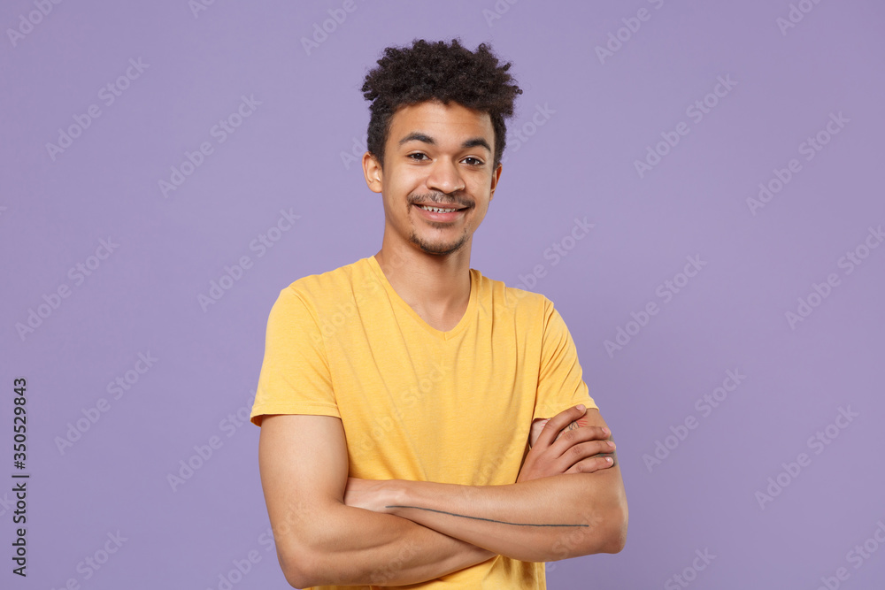 Obraz premium Smiling young african american guy in casual yellow t-shirt posing isolated on pastel violet background studio portrait. People emotions lifestyle concept. Mock up copy space. Holding hands crossed.