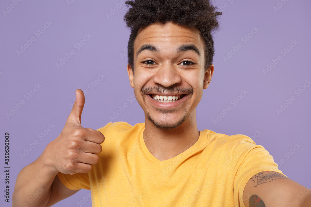 Close up of smiling young african american guy in casual yellow t-shirt posing isolated on pastel violet wall background. People lifestyle concept. Doing selfie shot on mobile phone, showing thumb up. Close up of smiling young african american guy in casual yellow t-shirt posing isolated on pastel violet wall background. People lifestyle concept. Doing selfie shot on mobile phone, showing thumb up.