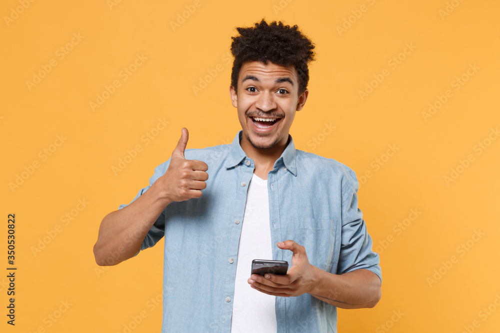 Cheerful young african american guy in casual blue shirt posing isolated on yellow wall background studio portrait. People lifestyle concept. Mock up copy space. Using mobile phone, showing thumb up. Cheerful young african american guy in casual blue shirt posing isolated on yellow wall background studio portrait. People lifestyle concept. Mock up copy space. Using mobile phone, showing thumb up.
