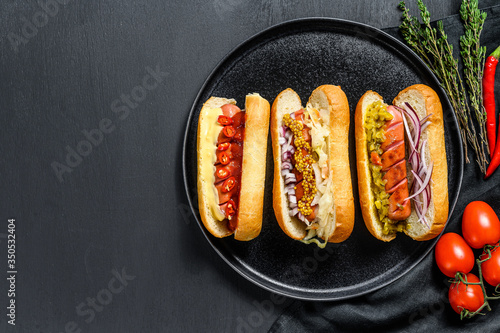 Hot dogs fully loaded with assorted toppings on a tray. Delicious hot-dogs with pork and beef sausages. Black background. Top view. Copy space