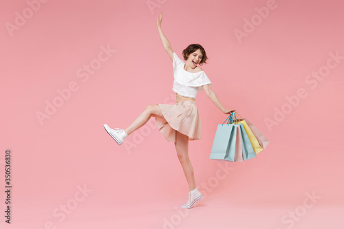 Funny young woman girl in summer clothes hold package bag with purchases isolated on pastel pink background. Shopping discount sale concept. Mock up copy space. Dancing, spreading hands, rising leg.