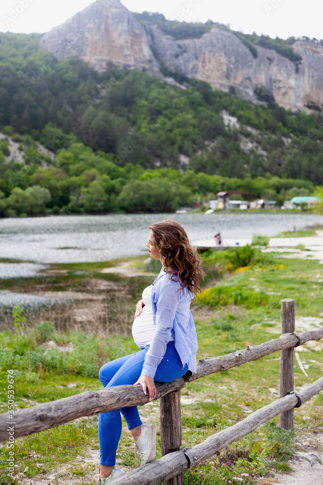 Naklejka premium Adult pregnant woman relaxing in the nature against the lake . Pretty girl looking to the tummy with love, female dressed blue jeans, white t-shirt, and shirt