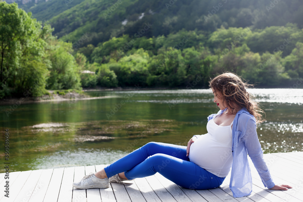 Adult pregnant woman relaxing in the nature against the lake . Pretty girl looking to the tummy  with love, female dressed blue jeans, white t-shirt, and shirt