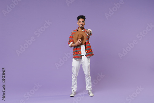 Smiling young african american guy in casual colorful shirt posing isolated on violet background studio portrait. People lifestyle concept. Mock up copy space. Hold hat, point index finger on camera.