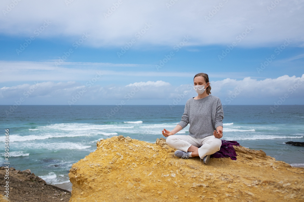 Peaceful woman in medical mask, practicing yoga on beach, meditating in lotus pose, keeping arms in zen gesture. High angle, copy space. Life balance during outbreak concept