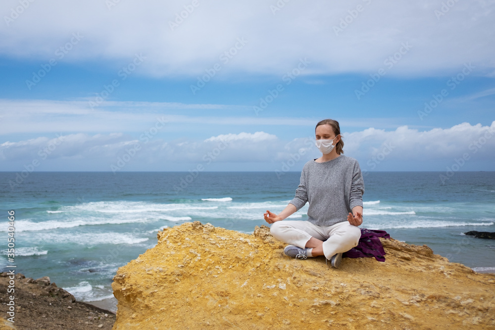 Peaceful woman in medical mask, practicing yoga on beach, meditating in lotus pose, keeping arms in zen gesture. High angle, copy space. Life balance during outbreak concept Peaceful woman in medical mask, practicing yoga on beach, meditating in lotus pose, keeping arms in zen gesture. High angle, copy space. Life balance during outbreak concept