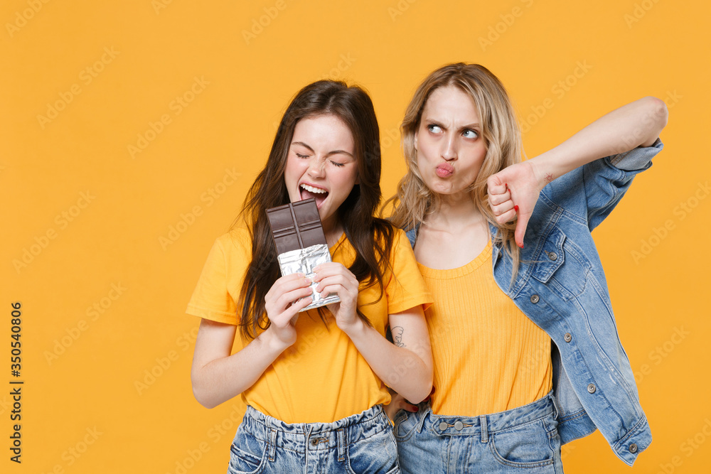Two confused young women girls friends in casual t-shirts denim clothes isolated on yellow background studio. People lifestyle concept. Mock up copy space. Biting chocolate bar, showing thumb down.