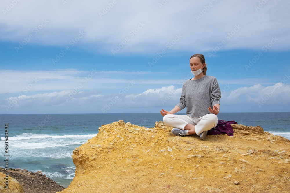 Calm young woman in face mask meditating at ocean, keeping arms in zen gesture. Copy space, low angle. Staying well during outbreak concept Calm young woman in face mask meditating at ocean, keeping arms in zen gesture. Copy space, low angle. Staying well during outbreak concept