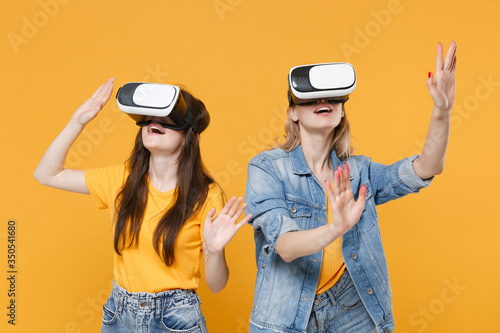 Women girls friends in casual t-shirts denim clothes isolated on yellow background. People lifestyle concept. Watching in headset  touch something like push on button point at floating virtual screen.