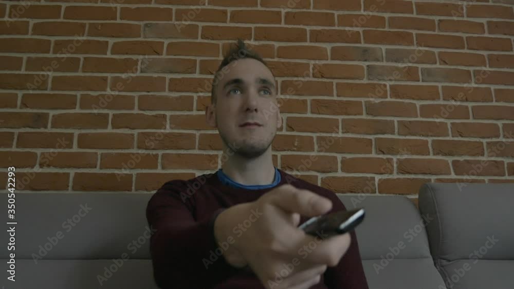 A man sitting on a couch and pointing at the TV with his remote control. He switches one program after another and looks for something he will enjoy. Bored expression A man sitting on a couch and pointing at the TV with his remote control. He switches one program after another and looks for something he will enjoy. Bored expression