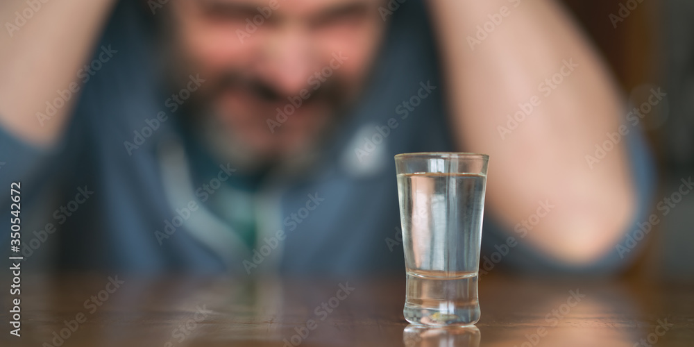 depressed man clutches his head with hands sitting at table with glass of spitrt