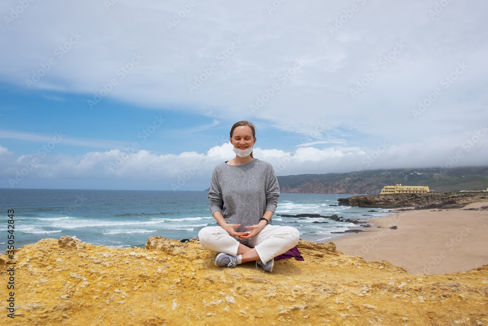 Positive young woman with closed eyes in face mask, practicing yoga and meditation on ocean beach. Front view. Staying well during outbreak concept