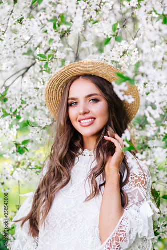 Portrait of young smiling woman in straw hat in the flowered garden in the spring time. © Elena Kratovich