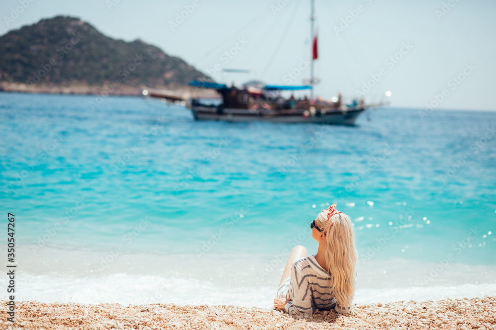 Young blonde woman relaxing on the beach. Summer lifestyle portrait of pretty sexy girl. Enjoying life and sitting on the beach of the island. looks into the distance at a sailing ship on the horizon.