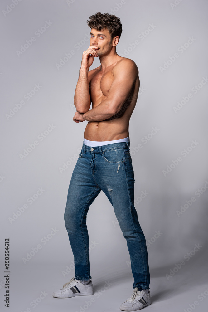 muscular man in denim jeans looking at camera and touching face on grey muscular man in denim jeans looking at camera and touching face on grey