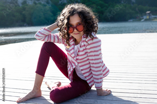 Pretty girl dressed red jeans , sunglasses and white t-shirt relax in the nature alone against the beautiful view of mountain, forest, sunrise and lake.