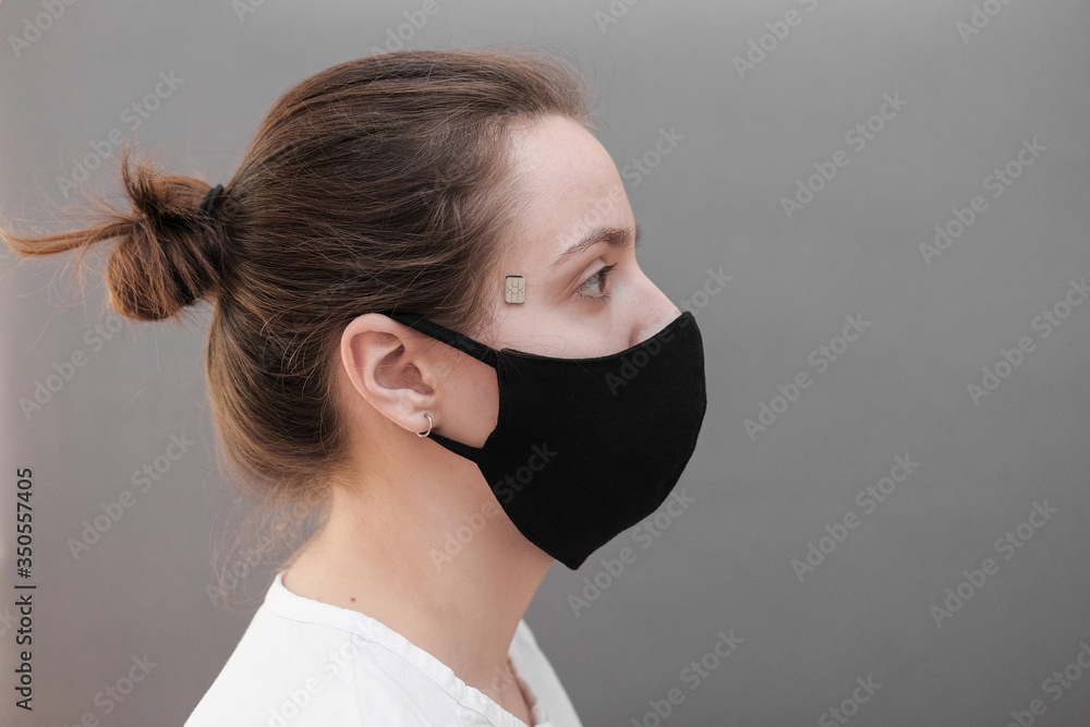 Chipization of people. A microchip implanted on a person's temple during a coronavirus disease to monitor life indicators. A woman in a black protective medical mask. Chipization of people. A microchip implanted on a person's temple during a coronavirus disease to monitor life indicators. A woman in a black protective medical mask.