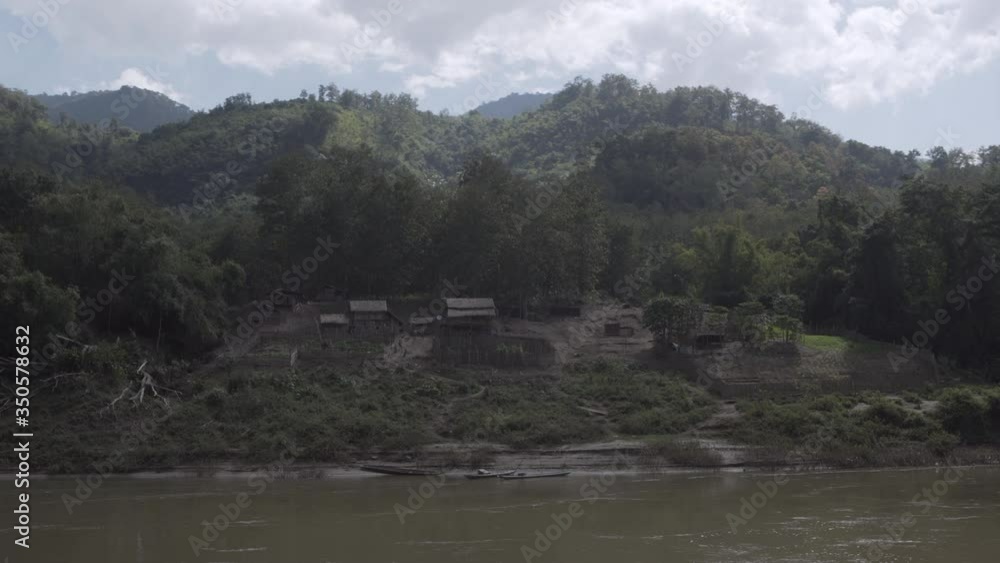 Panning shot of structures by trees at riverbank against mountains - Luang Prabang, Laos Panning shot of structures by trees at riverbank against mountains - Luang Prabang, Laos