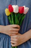 Front view of a woman wearing a blue jean dress, holding a bouquet of artificial tulips in red, yellow, pink, and white color with both hands. Feeling the love, warmth, and hope. Special day concept.