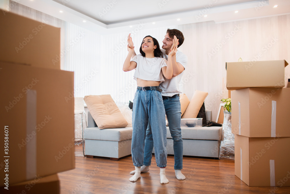 A young married couple in the living room in the house stand near unpacked boxes. hey are happy about new home. Moving, buying a house, apartment concept. A young married couple in the living room in the house stand near unpacked boxes. hey are happy about new home. Moving, buying a house, apartment concept.