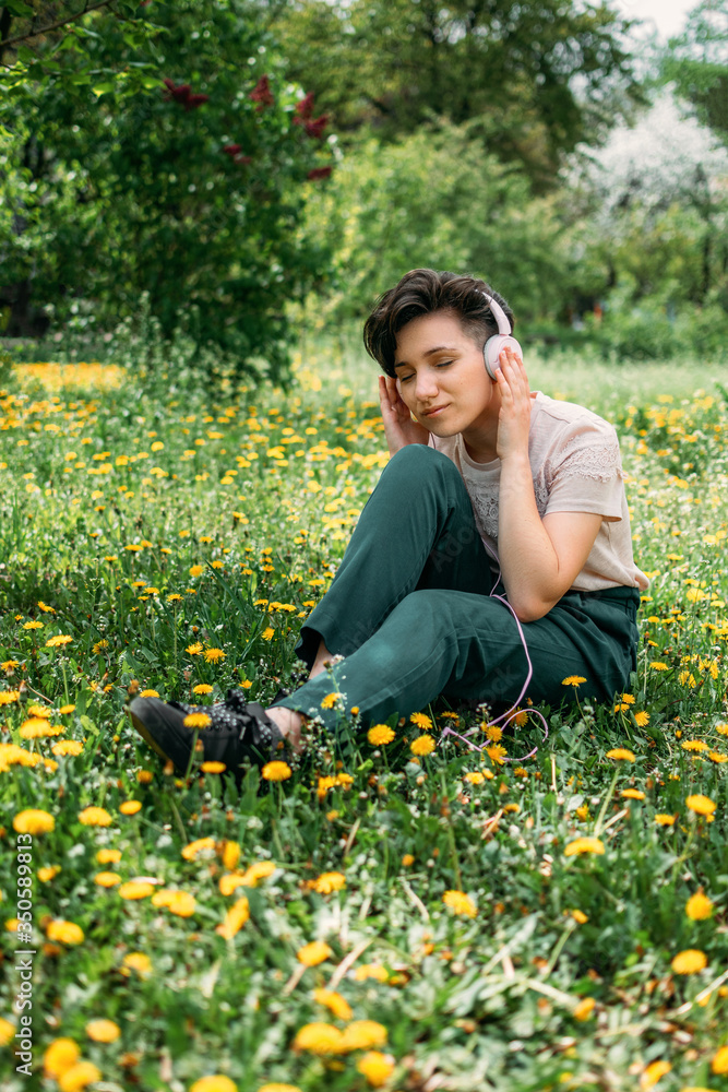 Young beautiful woman sitting on the lawn green grass with dandelions and listen to music in headphones. Young gen z girl listening to music on headphones in a summer park Young beautiful woman sitting on the lawn green grass with dandelions and listen to music in headphones. Young gen z girl listening to music on headphones in a summer park
