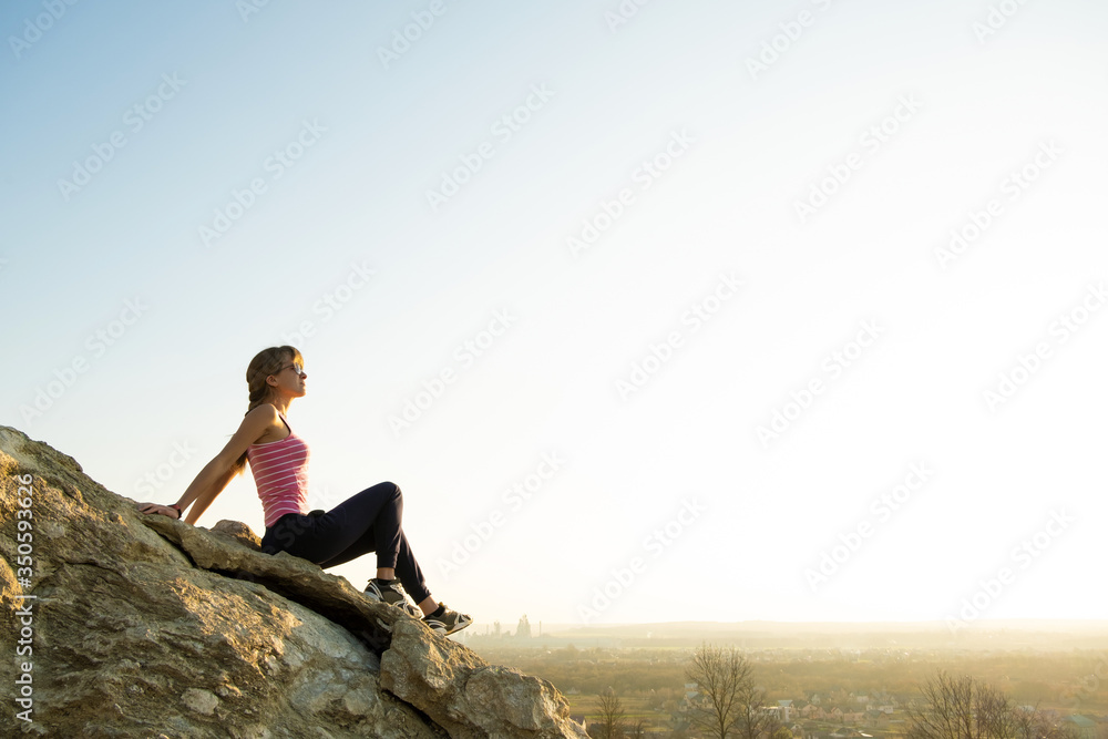 Woman hiker sitting on a steep big rock enjoying warm summer day. Young female climber resting during sports activity in nature. Active recreation in nature concept.