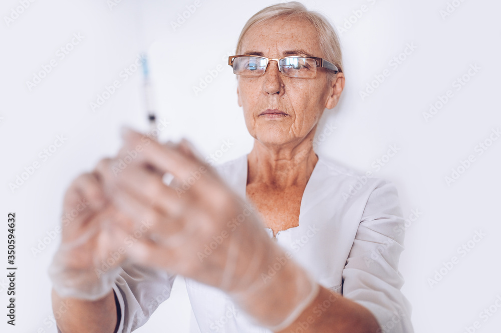 Elderly senior mature woman doctor or nurse with syringe in a white medical coat and gloves wearing personal protective equipment isolated. Healthcare and medicine concept. Covid-19 pandemic crisis Elderly senior mature woman doctor or nurse with syringe in a white medical coat and gloves wearing personal protective equipment isolated. Healthcare and medicine concept. Covid-19 pandemic crisis