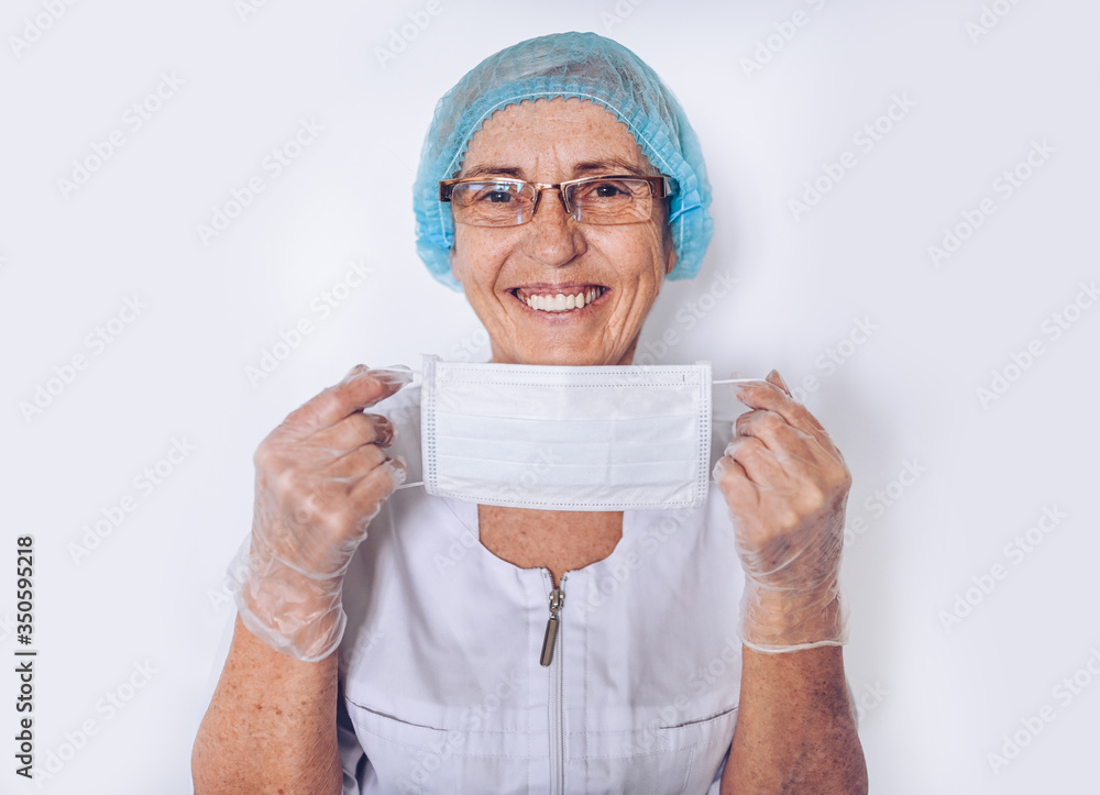Elderly smiling mature woman doctor or nurse in a white medical coat, gloves, puts on face mask wearing personal protective equipment isolated. Healthcare and medicine concept. Covid19 pandemic crisis Elderly smiling mature woman doctor or nurse in a white medical coat, gloves, puts on face mask wearing personal protective equipment isolated. Healthcare and medicine concept. Covid19 pandemic crisis