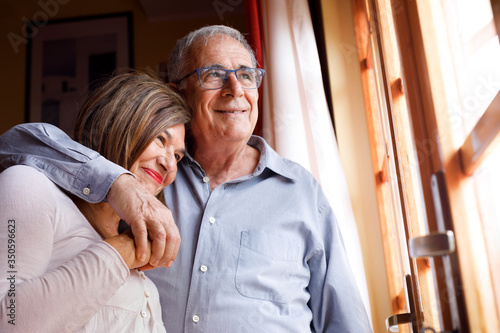 coppia di anziani si abbraccia felice e sorride in piedi nel salotto di casa photo