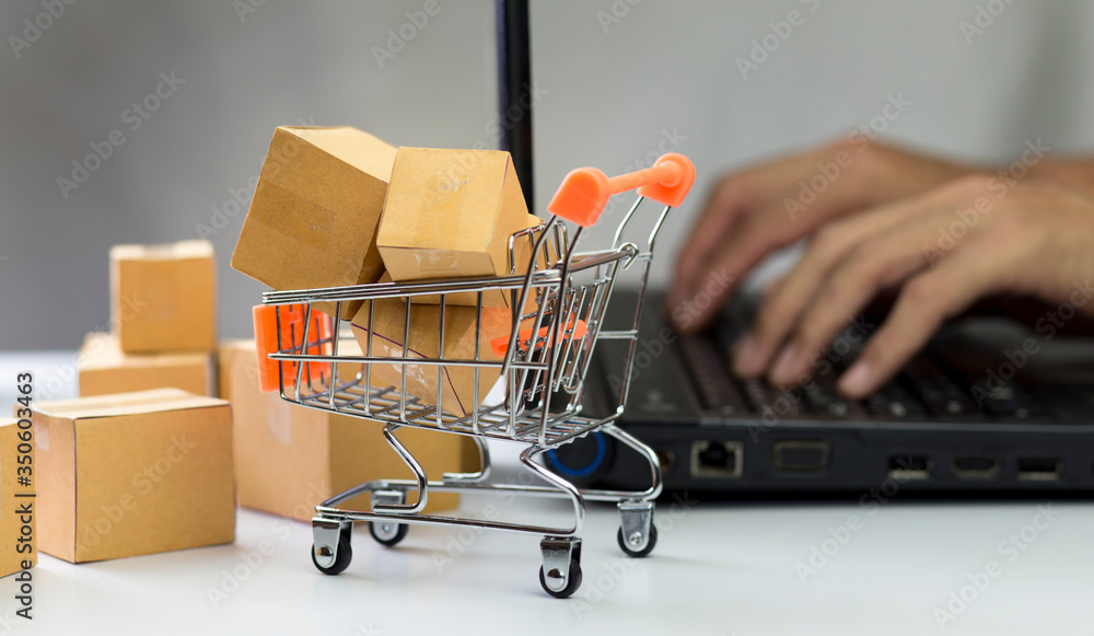 Supermarket shopping cart with loading cartons in and man hand using laptop background